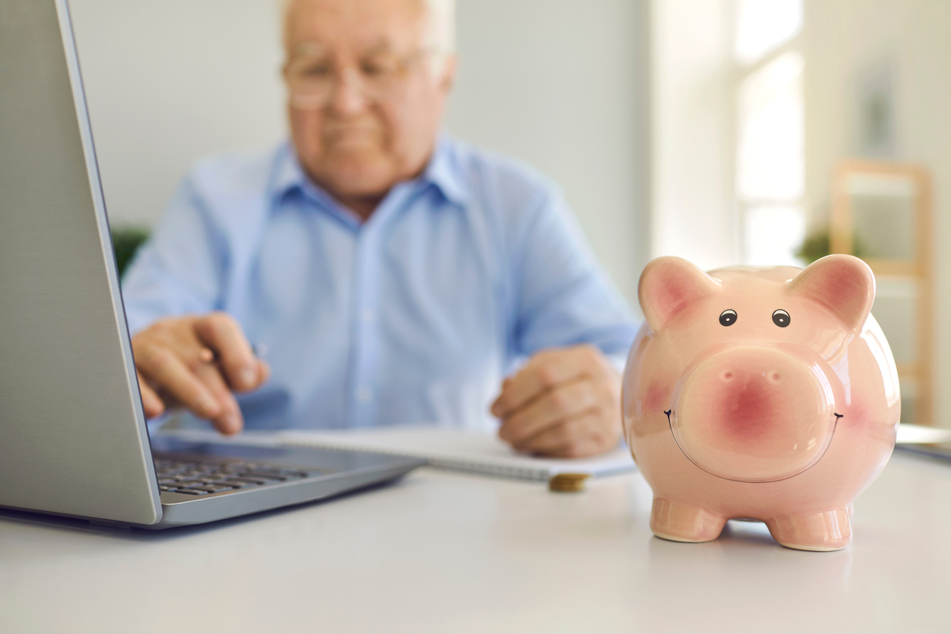 Piggy Bank Standing on Desk with Blurred Senior Man Using Laptop to Pay Bills Online in Background